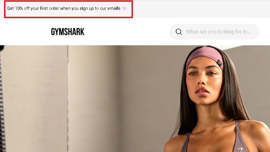 WordPress popup plugins: A banner at the top of the Gymshark website reads, Get 10% off your first order when you sign up to our emails, above a search bar and an image of a woman in workout clothing.