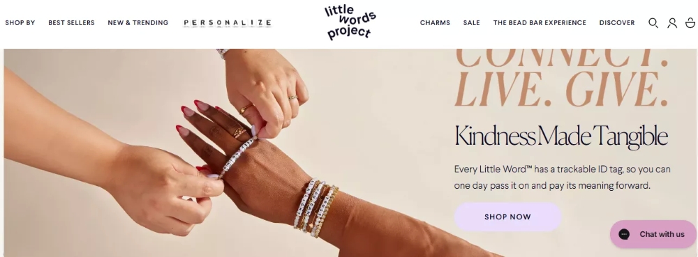 Omnisend vs. Shopify: Two hands fasten a beaded bracelet onto another persons wrist, with jewelry and bracelets visible. The text reads, “Kindness Made Tangible” and promotes tracking and sharing Little Words Project bracelets.