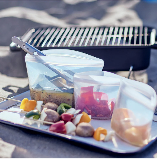 Most profitable products to sell online: A tray on sand holds veggie skewers, reusable silicone bags with marinating food, and tongs, with a portable grill in the background.