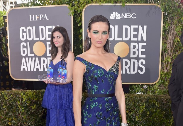 Guerilla marketing: A woman in a blue dress with green patterns poses confidently on the Golden Globe Awards red carpet, while another woman in blue behind her carries a tray with bottles of water. Event signs and greenery are visible in the background.