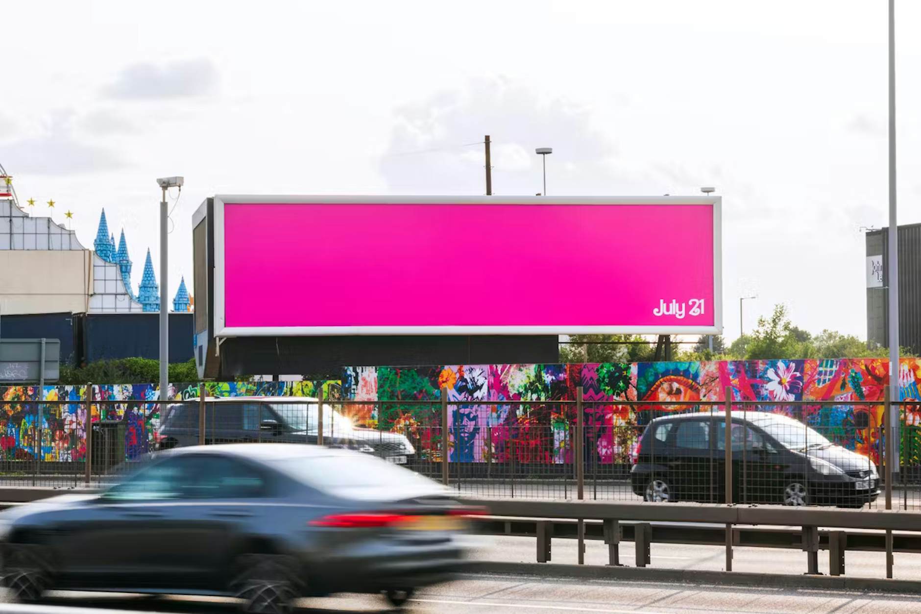 Guerilla marketing: A large, bright pink billboard with the text July 21 stands above a colorful graffiti-covered wall beside a busy road with blurred cars passing by.