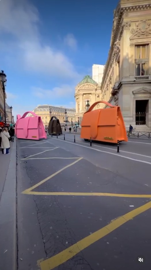Guerilla marketing: Large pink, brown, and orange handbags are being driven like cars down a city street lined with historic European-style buildings under a blue sky.
