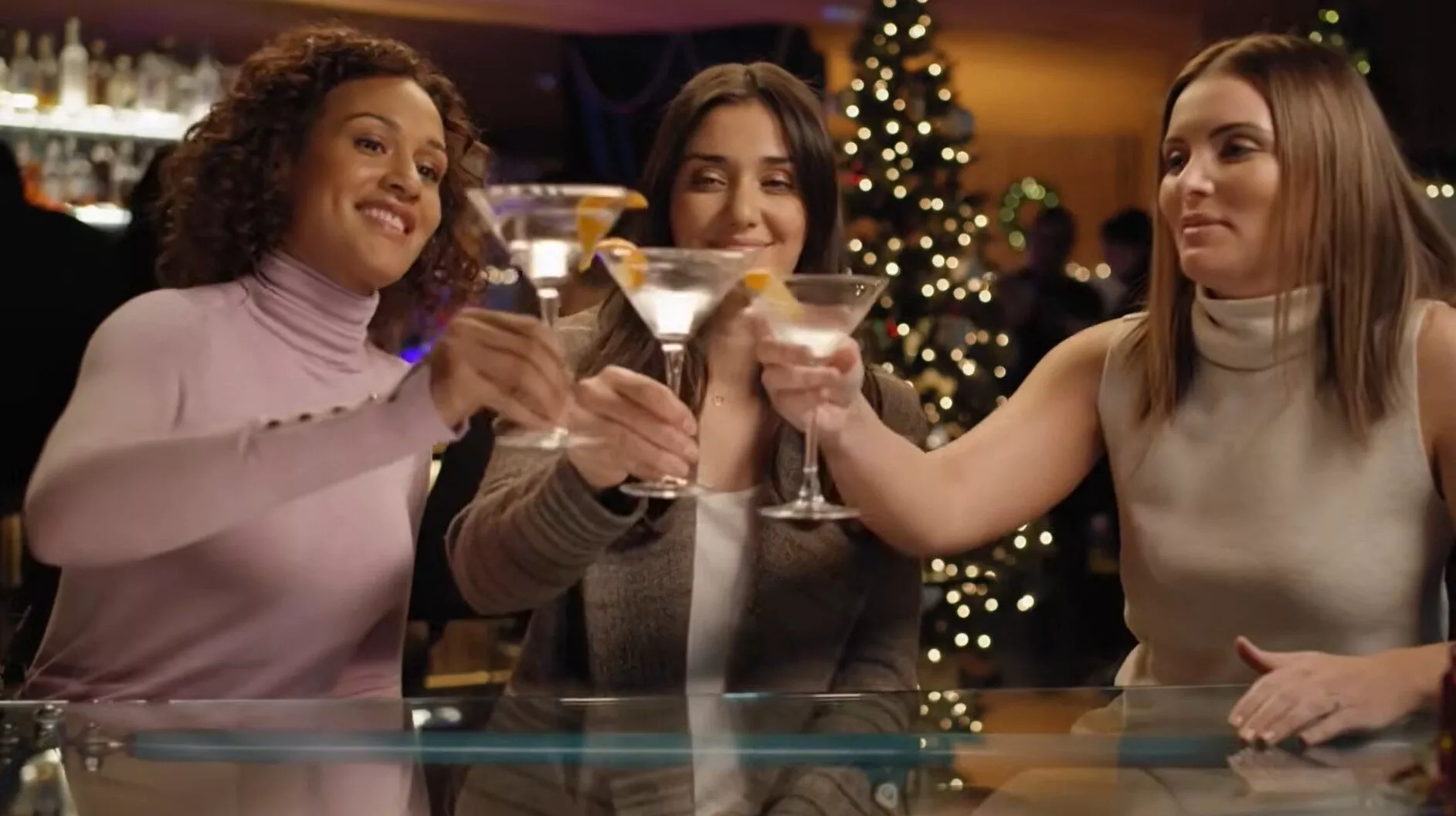 Guerilla marketing: Three women sitting at a bar, smiling and clinking glasses with cocktails. There are festive lights and a decorated Christmas tree in the background.