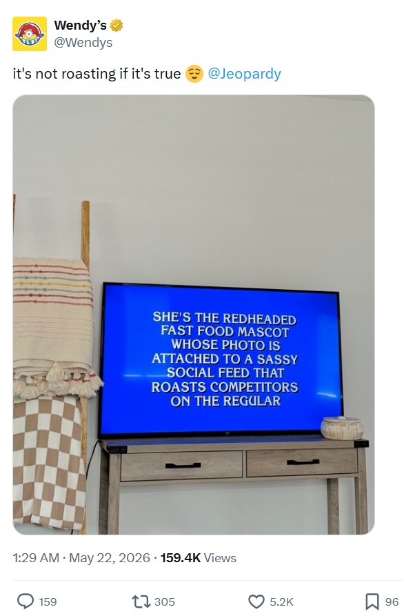 Guerilla marketing: A TV shows a Jeopardy! clue about a redheaded fast food mascot known for roasting competitors on social media. The TV is on a wooden stand beside a basket and a folded blanket.