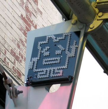 Guerilla marketing: A pixelated cartoon character made of small lights is mounted on a metal sign under a structure, with a brick wall in the background.