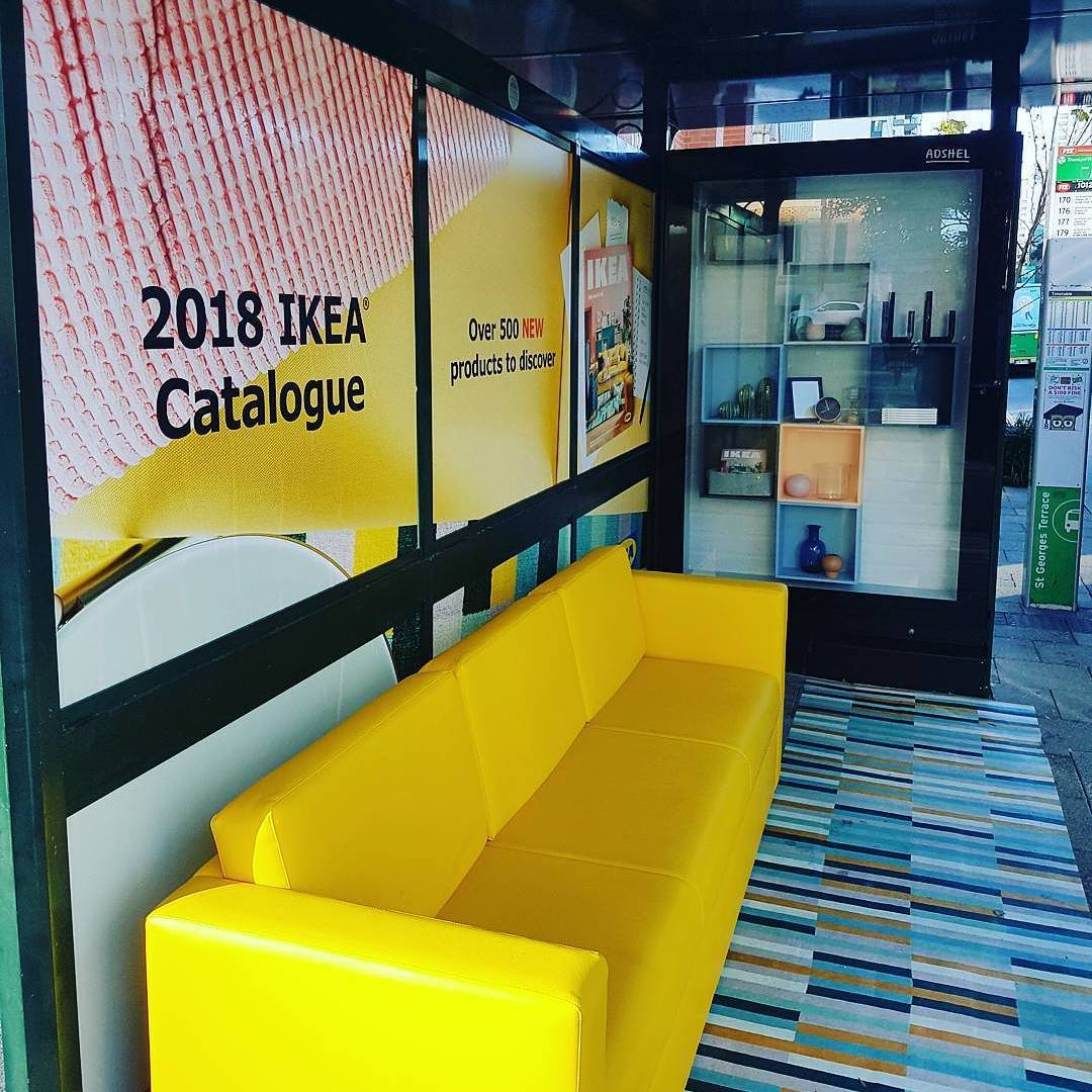 Guerilla marketing: A bus stop decorated like a living room, featuring a bright yellow sofa, a striped rug, a wall ad for the 2018 IKEA Catalogue, and a glass display case with shelves and various home items.