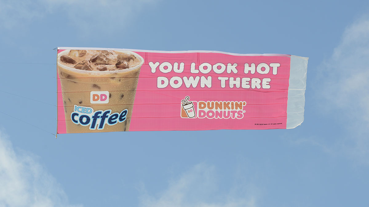 Guerilla marketing: A banner flying in the sky shows an iced coffee and the words YOU LOOK HOT DOWN THERE beside Dunkin’ Donuts branding and logo on a pink background.