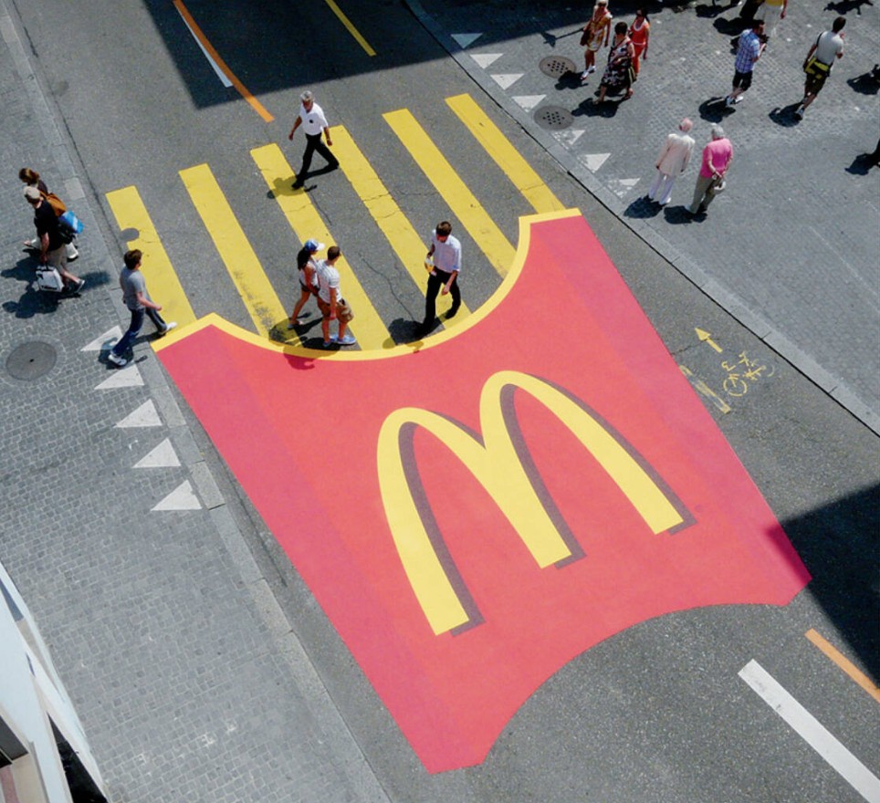 Guerilla marketing: A crosswalk painted to look like a red McDonald’s fries box with yellow fries serves as the crosswalk lines, with people walking across it on a city street.