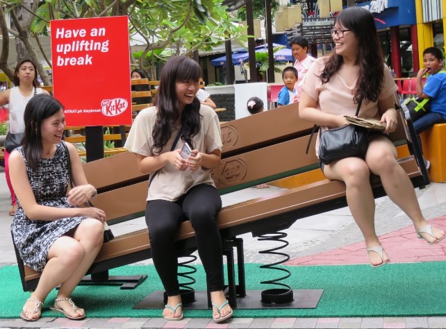 Guerilla marketing: Three women sit and laugh on a bench with springs causing one side to tilt upward. A red KitKat sign behind them reads Have an uplifting break. Several people watch in the background.