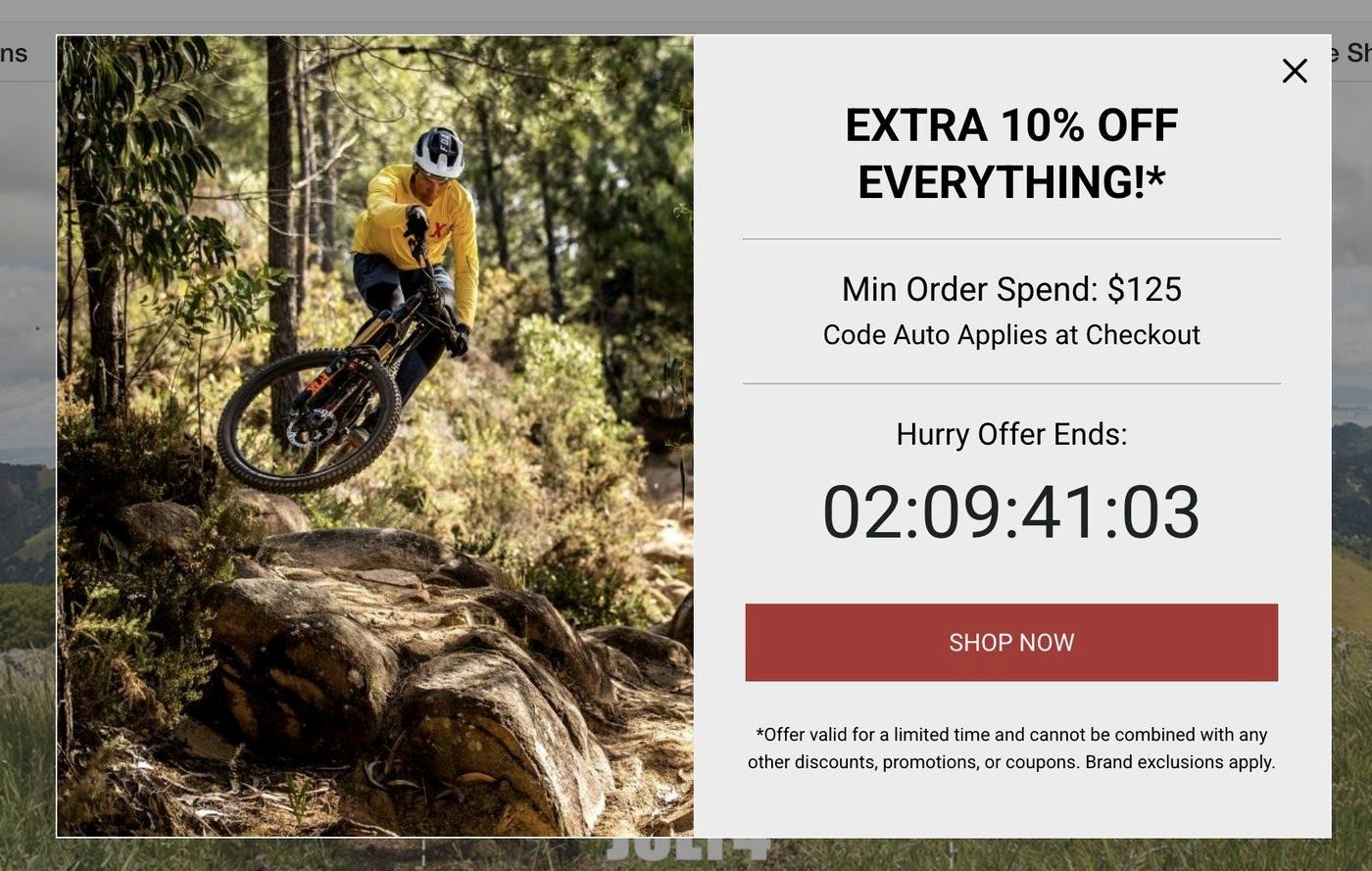 Black friday popup: A person mountain biking over rocks in a forest. Next to the rider, a promo box offers Extra 10% Off Everything with a $125 minimum spend, a countdown timer, and a red Shop Now button.