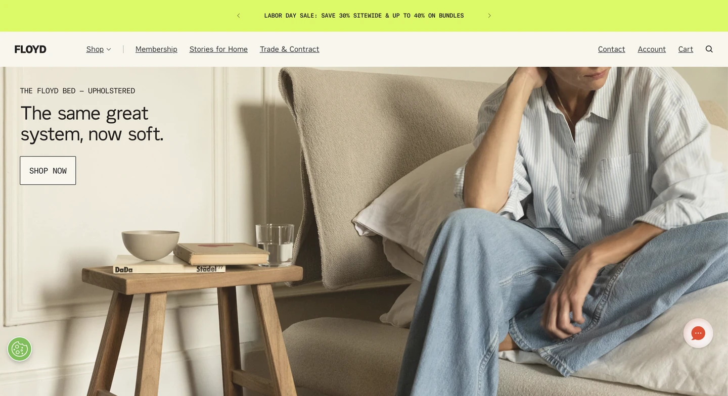 Person in a striped shirt and jeans sits on a beige sofa next to a wooden side table with books, water, and glasses.