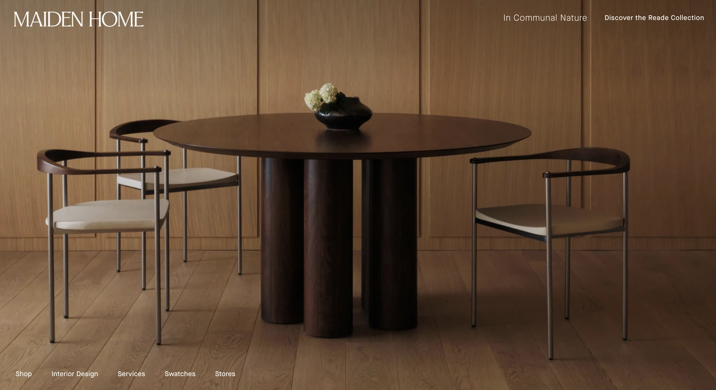 Round wooden dining table with four modern chairs in a minimal room with wood-paneled walls and light wood flooring.