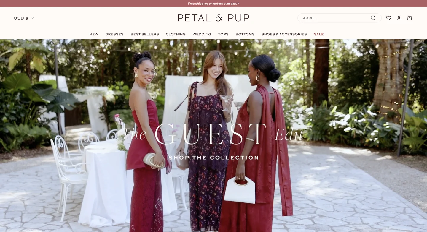 Three women in formal dresses stand outdoors near a white table, smiling and talking.