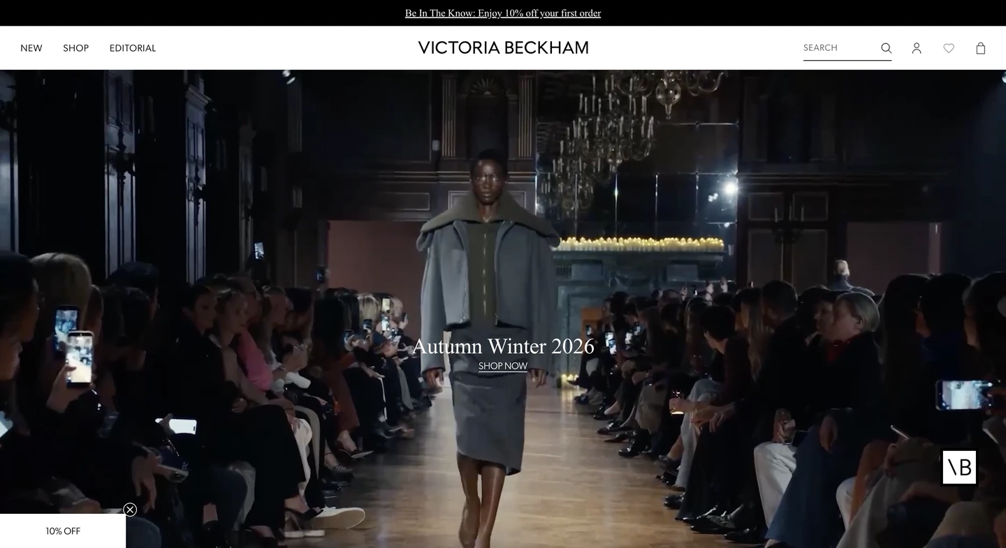 Model walks down a runway in a gray outfit at a fashion show, with audience seated on both sides.
