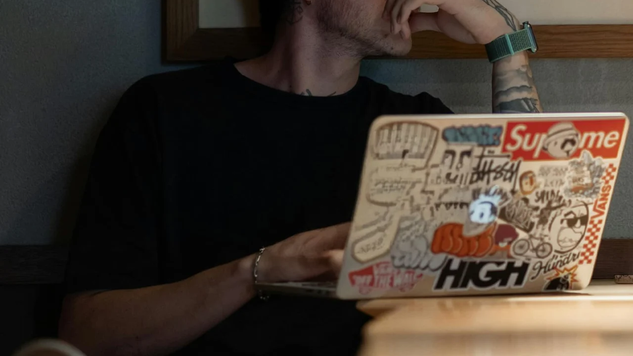 A person in a black t-shirt sitting at a wooden table with a sticker-covered laptop open on their lap, hand resting against their chin.